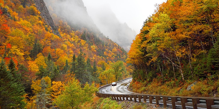 Foggy mountains full of fall foliage while a car drives through on an intersecting road at the Moose Path Trail