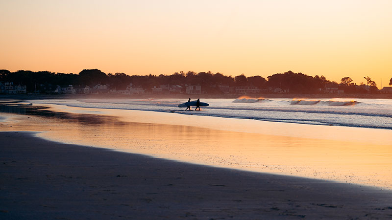 a surfer walking down a beach at sunset